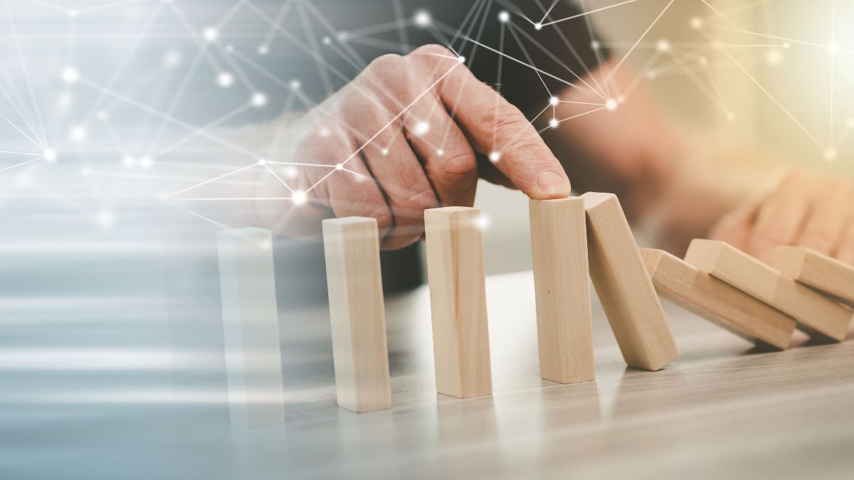 Eine Hand stößt mit dem Zeigefinger in einer Reihe aufgestellte Holzblöcke um. Darüber ist eine digitale Illustration von mit Linien verbundenen Punkten zu sehen.